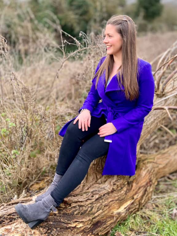 Cambridgeshire Contemporary Celebrant, Jemma in a long blue coat with straight brown hair. Jemma is sat on a log smiling and excited to start planning your ceremony.